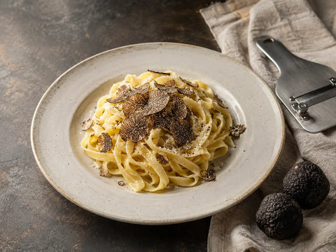 Tagliatelle fekete szarvasgomba-forgáccsal, egész szarvasgombákkal és szarvasgombaszeletelővel egy kő munkalapon.